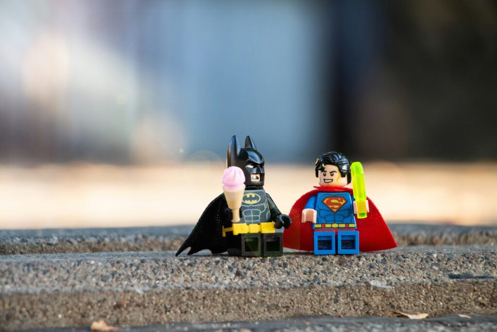 Two Lego figures, one as Batman holding an ice cream cone and the other as Superman with a popsicle, stand on a pavement with a blurred background.