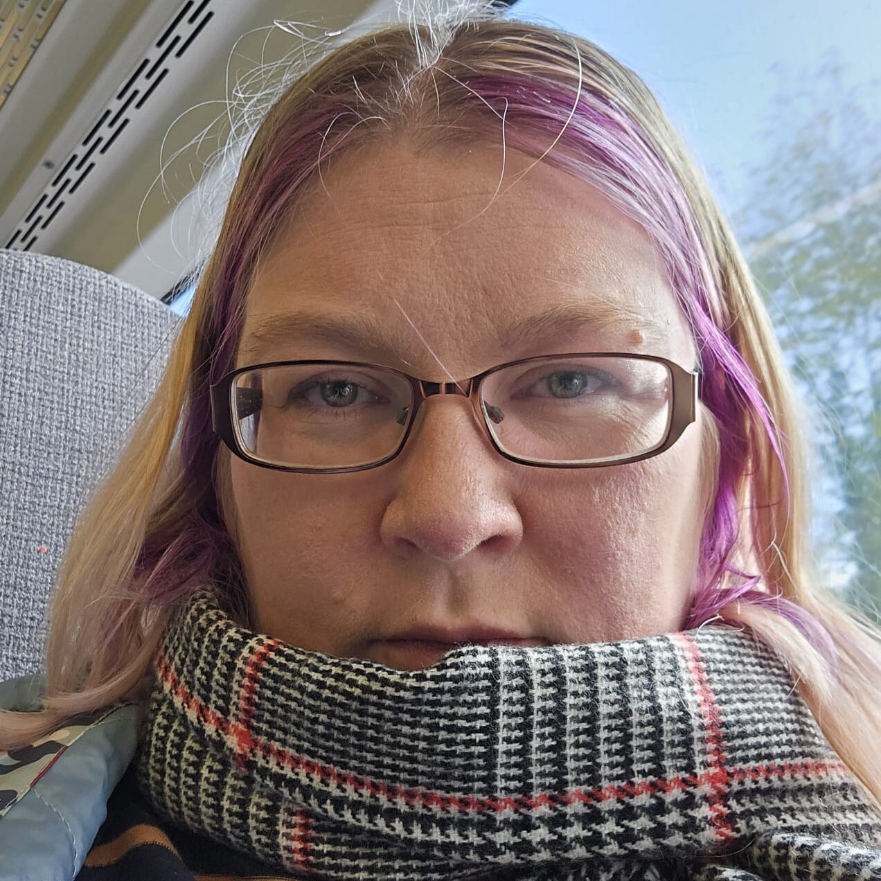 Katie Stone, with purple hair tips and glasses, sitting by a window on a train, wrapped in a plaid scarf.