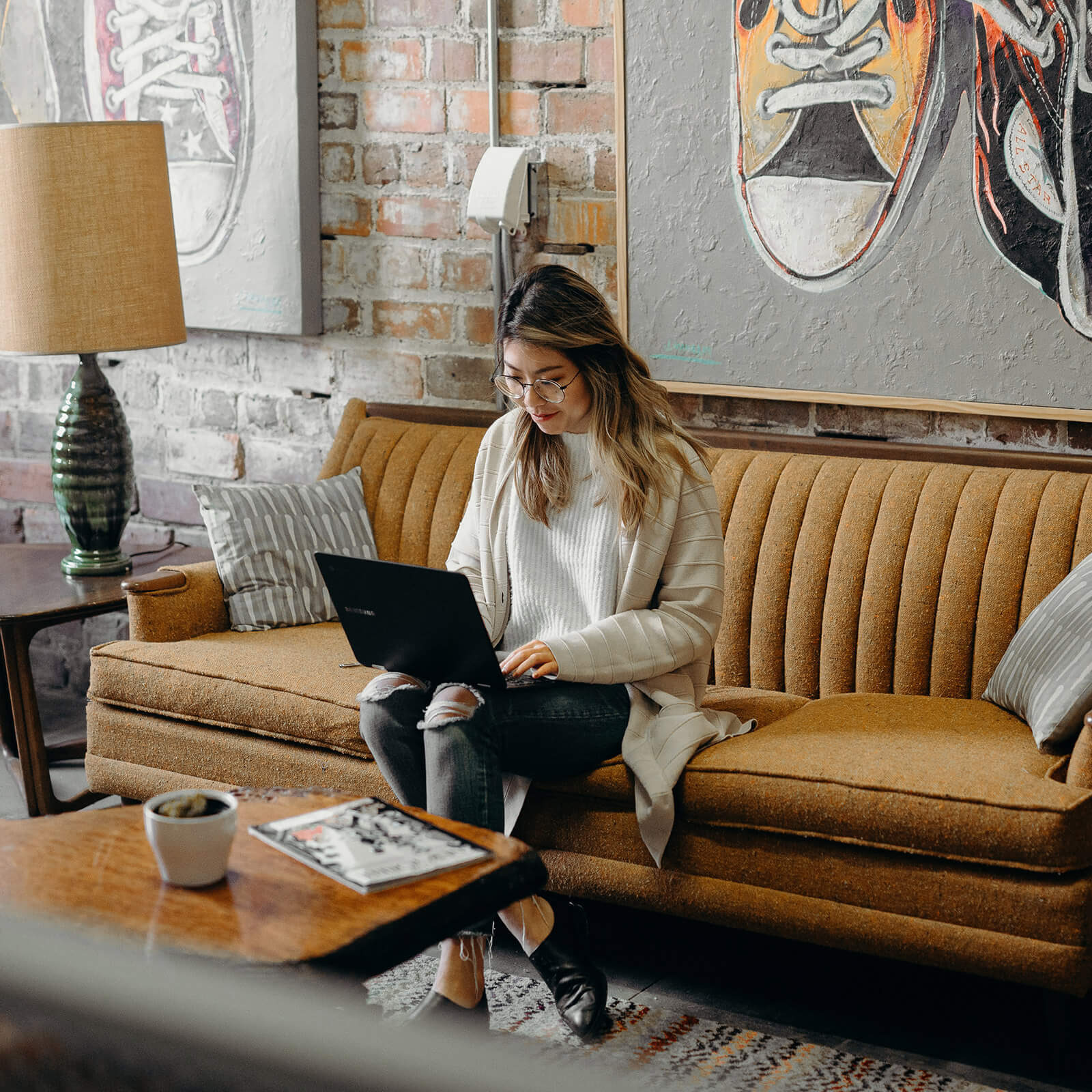Woman sitting on a brown sofa using a laptop in a stylish living room with brick walls, modern art, and warm lighting—perfect for an efficient UK based virtual assistant at work.